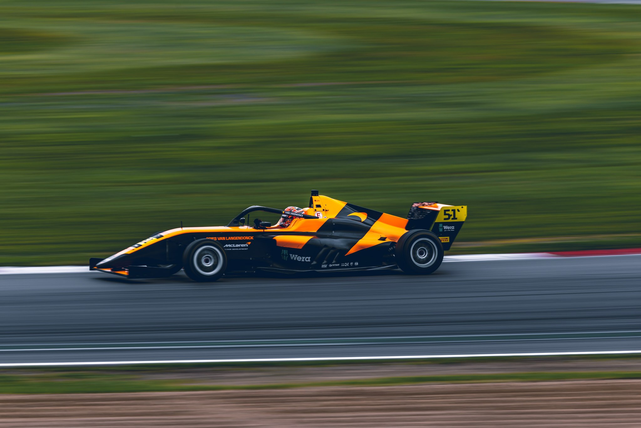 Orange and black number 51 single-seater moving across the frame against a motion-blurred track and grass.